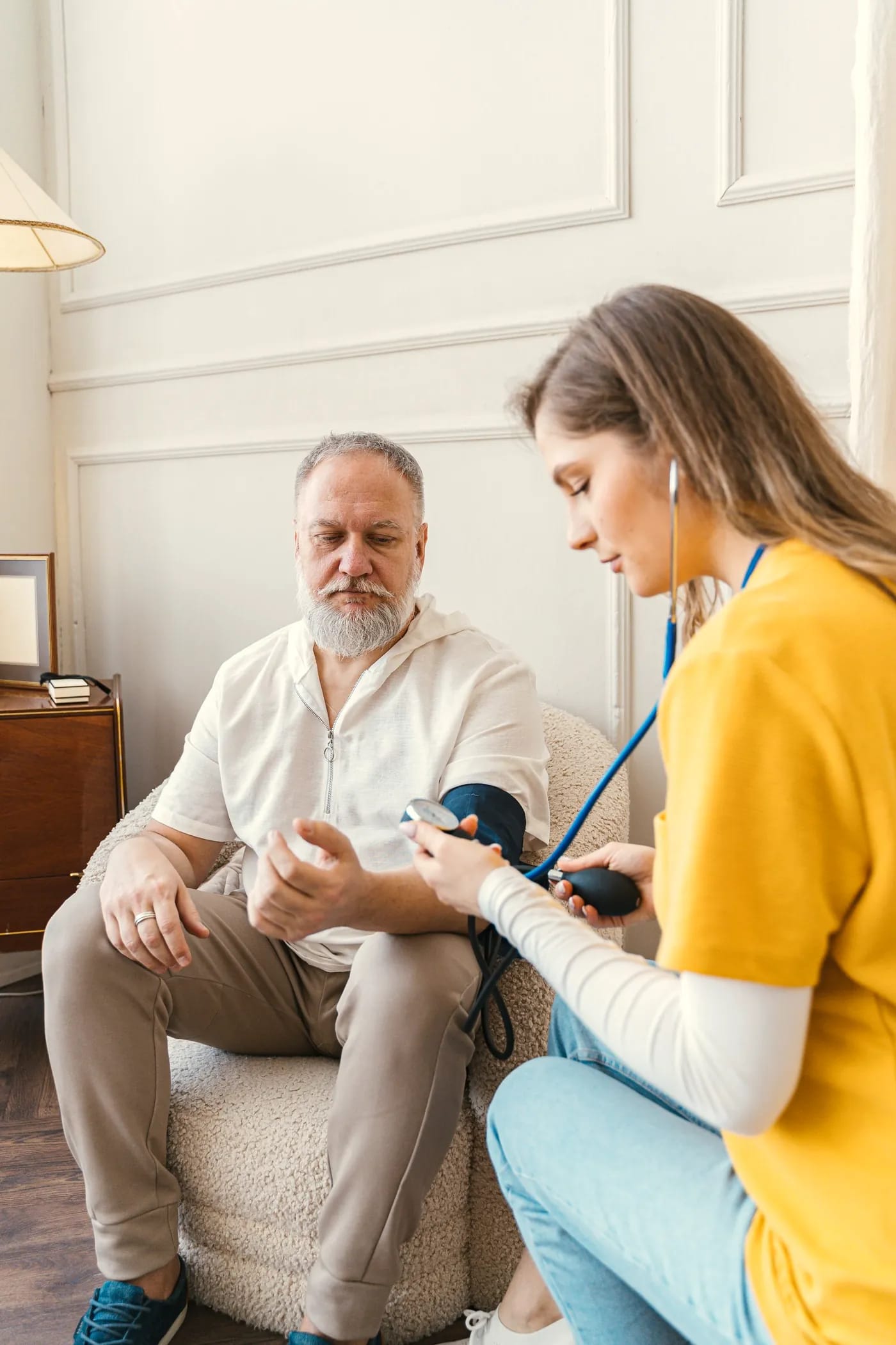 A hospice nurse takes an older man’s blood pressure while he sits in an armchair at home.
